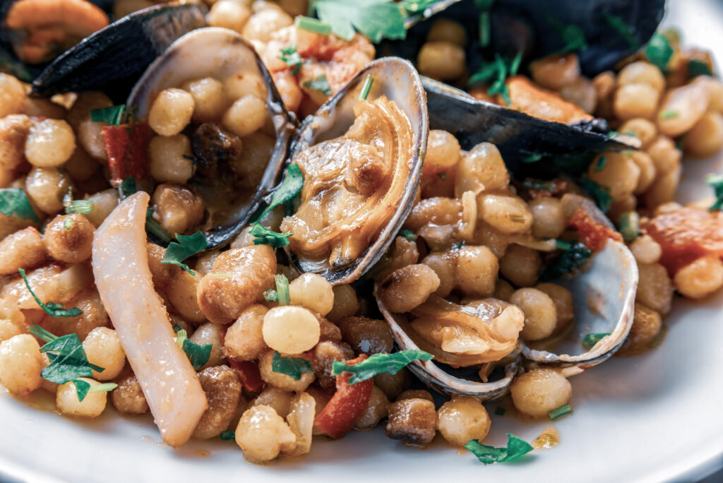 Fregola Sarda mit Meeresfrüchten und Muscheln auf einem weißen Teller, angerichtet mit frischer Petersilie