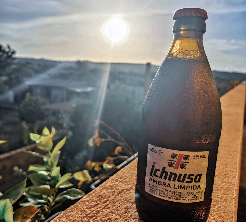Eine Flasch Ichnusa Bier Sardinien, das auf dem Balkongeländer steht und im Hintergrund der sardische Sonnenuntergang