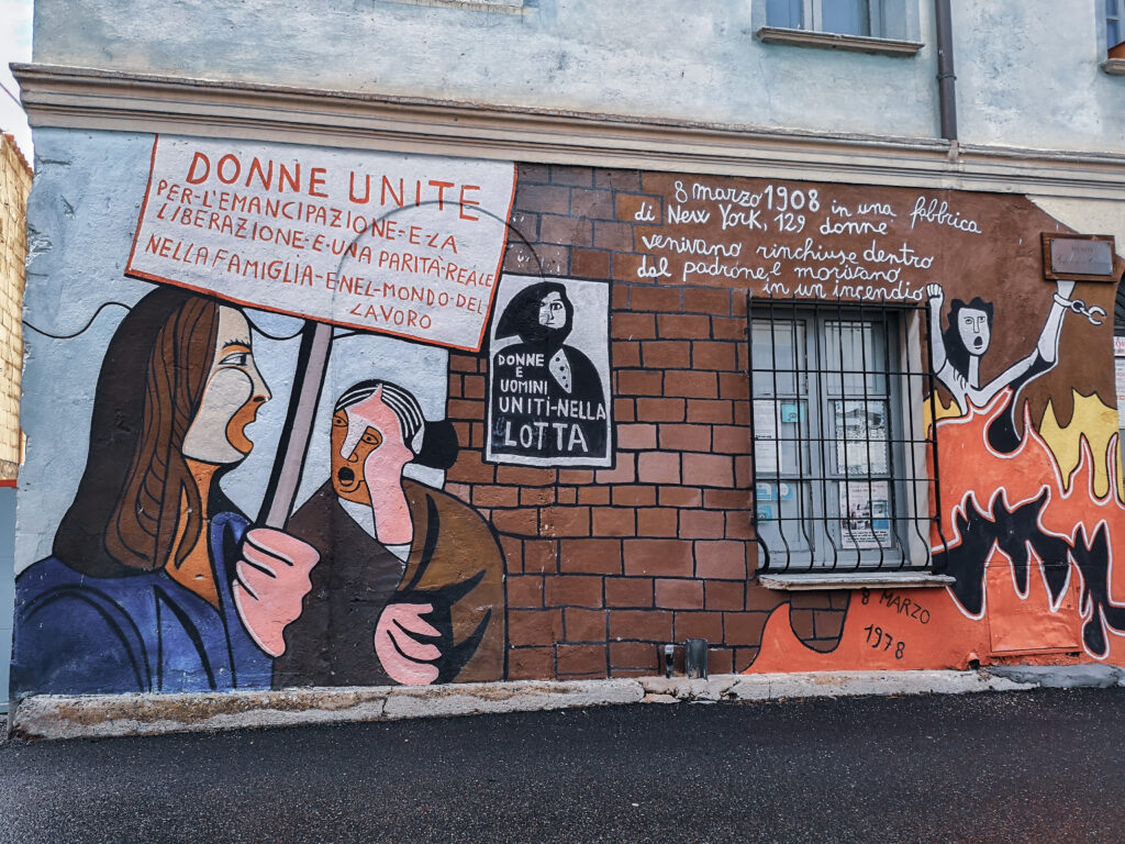 Murales in Orgosolo mit Frauenprotest, Flammen und historischem Verweis auf 129 getötete Arbeiterinnen im Jahr 1908 in New York
