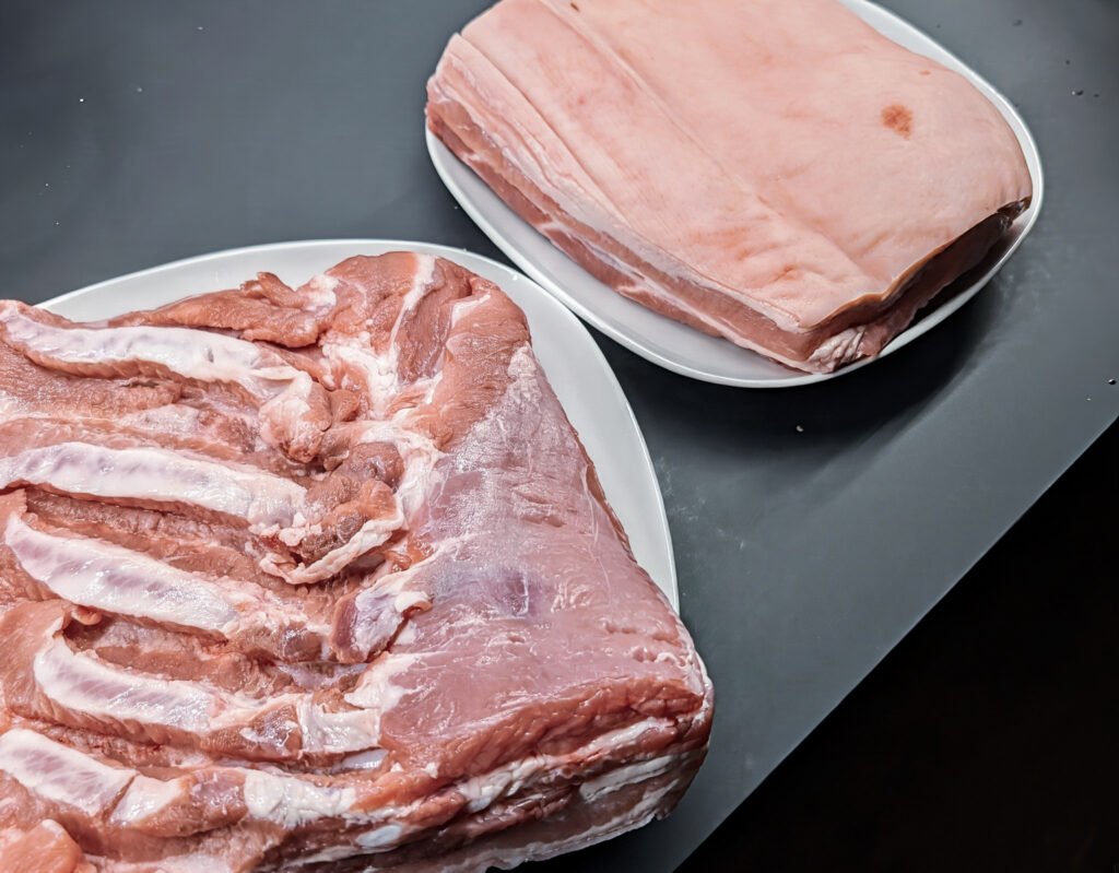 Zwei rohe Schweinebauchstücke auf Tellern, eines mit sichtbarer Fleischseite und eines mit heller Schwarte
