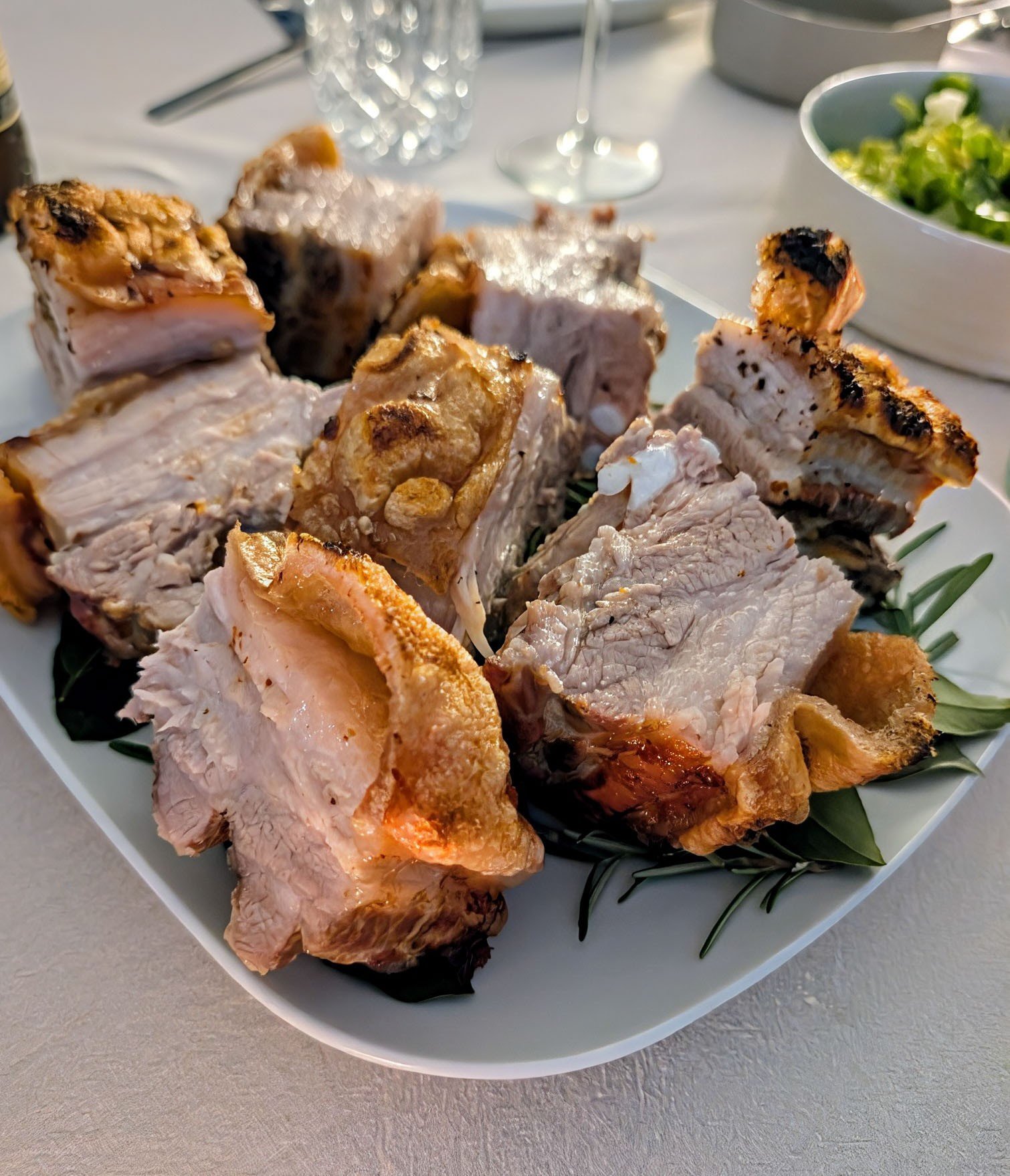 Mehrere Stücke gebratener Schweinebauch mit krosser Schwarte auf einer weißen Servierplatte mit Kräutern